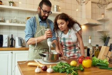 Italian man, chef cook using hand blender while preparing a meal. Young woman, girlfriend in apron looking at the process, helping in the kitchen