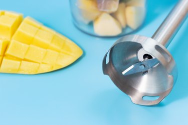 Immersion blender and mango on blue background close-up.