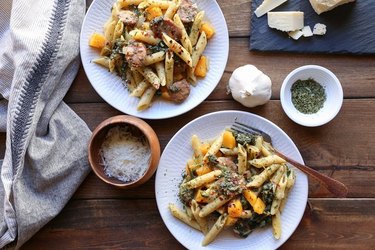 Overhead shot of two white plates of penne with squash and sausage on a rustic table, surrounded by the dish\'s ingredients and a clean kitchen towel