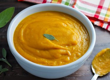 A white bowl of squash soup, garnished with a single sage leaf, surrounded by a spoon, napkin and additional sage leaves