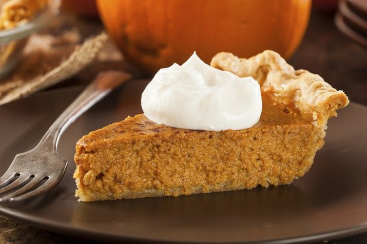 A slice of pumpkin pie, topped with whipped cream, on a dark plate with a whole pumpkin in the background