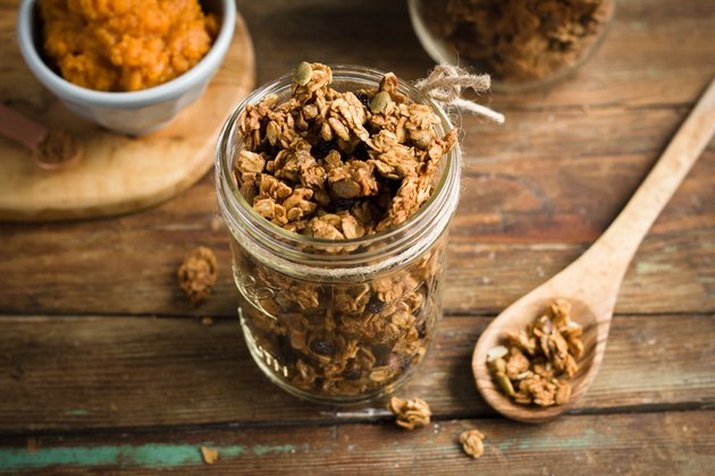 A mason jar full of pumpkin spice granola on a rustic table, with a wooden mixing spoon and a small bowl of pumpkin puree on either side